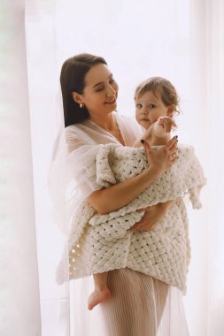 Beyaz elbiseli anne pencerenin yanında duruyor ve bebeğini kollarına alıp örgü örmüş bir battaniyeye sarılıyor. Ebeveyn koruması, bakım, sevgi, çocuk ve bebek sağlığı. Mutlu çocukluk kavramı. Metin için alanı kopyala