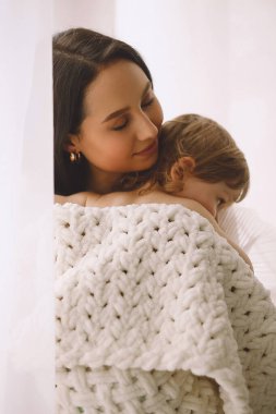 Mutlu anne, küçük kızına evde sarılıyor. Evde örgü örülü battaniyeye sarılı küçük bir kız. Ebeveyn koruması, bakım ve sevgi. Anne ve kızı.
