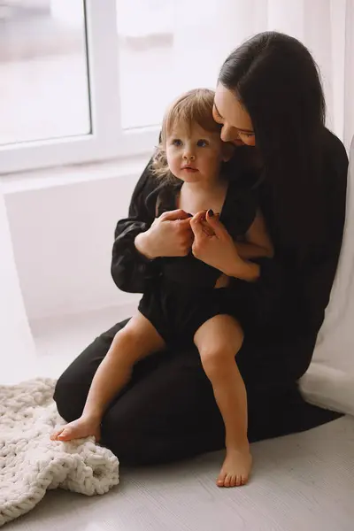 Mother and her two year old daughter in black outfits sitting near window in the studio and smiling. Happy mother hugging her little baby girl at home. Taking care of daughter. Daughters Day