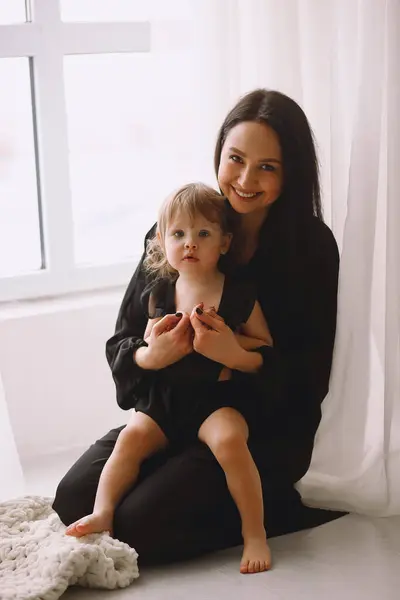 Mother and her two year old daughter in black outfits sitting near window in the studio and smiling. Happy mother hugging her little baby girl at home. Mother and daughter