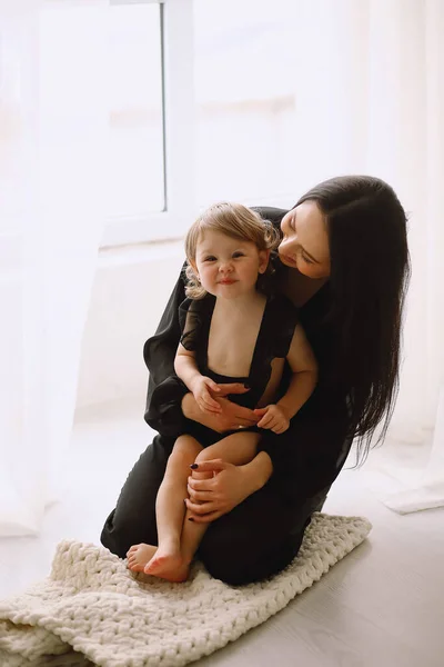Mother and her two year old daughter in black outfits sitting near window in the studio and smiling. Happy mother hugging her little baby girl at home. Mother and daughter