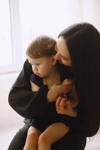 Mother and her two year old daughter in black outfits sitting near window in the studio. Happy mother hugging her little baby girl at home. Mother and daughter