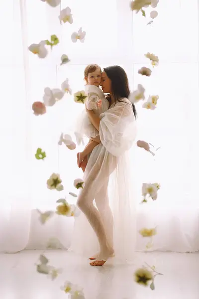 Studio portrait of happy mother in white dress standing near big window and hugging  her baby daughter. Mother and daughter in white outfit. The concept of happy motherhood