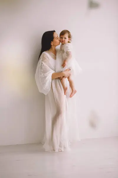 Happy mother in white outfit standing near wall in the studio and holding her baby daughter in her arms and kissing. Mother and daughter in white dresses. The concept of happy childhood. Copy space for text