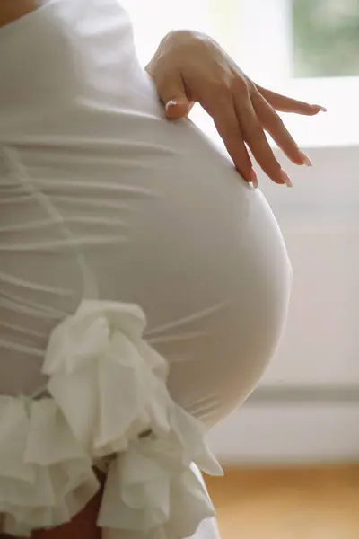 Close up photo of pregnant woman in a stylish white dress standing near window and touching her belly.  Stylish Motherhood concept. Maternity photoshooting