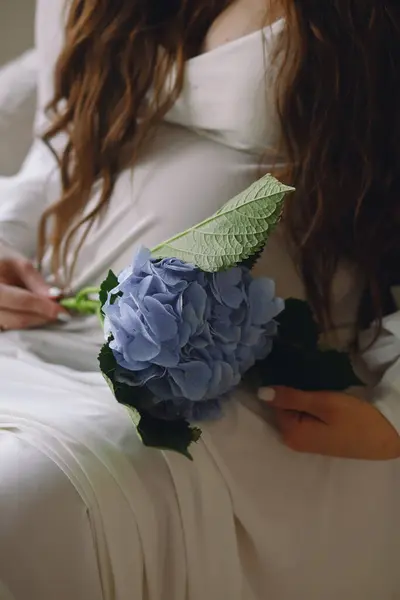 Pregnant brunette woman in a stylish white dress holding her belly with one hand and a blue hydrangea flower in the other hand. Close up photo of pregnant belly. Stylish Motherhood concept
