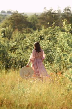 Pembe uçan elbisesi ve sol elinde hasır şapkası olan çekici esmer kadın. Genç bir kadın yazın doğada yürüyor. Güzellik ve moda. 