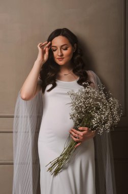 Beyaz elbiseli çekici hamile kadın elinde bir buket çigila tutuyor. Hamilelik fotosistemi. Mutlu annelik ve ebeveynlik kavramı.