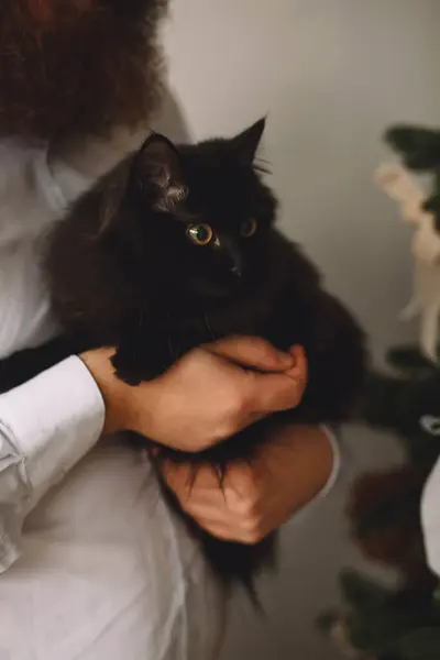 Man in white shirt holding his black cat in his hand at home. Owner and cat. Pet lovers. Family concept