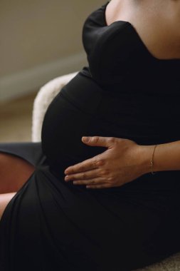 Kapalı bir fotoğrafta, klasik siyah elbiseli hamile bir kadın evinde sandalyeye oturmuş ve karnını tutuyordu. Tarz sahibi annelik kavramı. Doğum fotosop 'laması