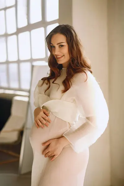 Portrait of smiling pregnant woman in stylish ivory dress in the studio. Stylish pregnancy concept. Beauty and fashion