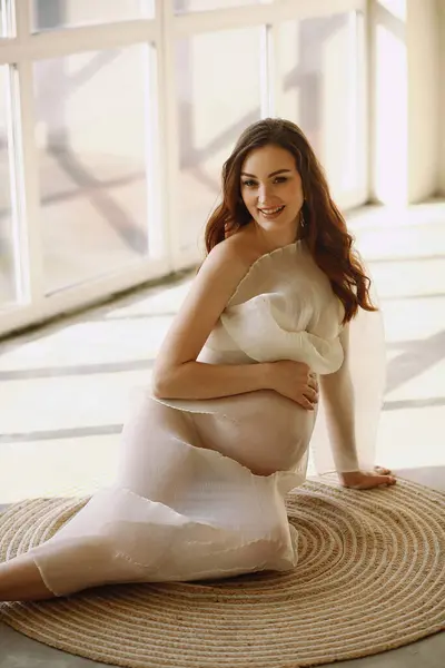Happy pregnant woman in stylish transparent dress sitting on the floor and posing in the studio. Stylish pregnancy