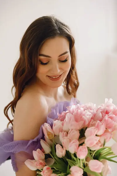 Attractive brunette woman in violet dress holding a pink bouquet of tulips while posing in the studio. Beauty and fashion. Copy space for text