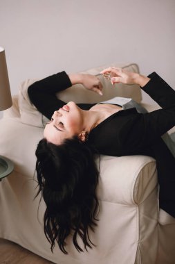 Siyah ceketli esmer kadın ve mavi gecelik sandalyede uzanıyor ve stüdyoda poz verirken bir şişe parfüm kullanıyor. Güzellik ve moda. Güzel genç bayan evde bir şişe parfüm kullanıyor. Metin için boşluk