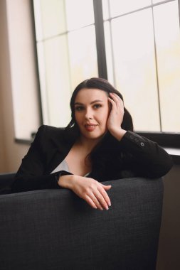 Siyah ceketli ve mavi gecelikli çekici esmer kadın pencerenin yanındaki sandalyede oturuyor ve stüdyoda poz veriyor. İş kadını. Güzellik ve moda