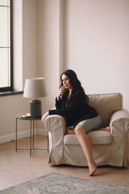 Siyah ceketli ve mavi gecelikli güzel esmer kadın sandalyede oturuyor ve stüdyoda poz verirken elinde parfüm tutuyordu. Uzayı metin için kopyala. Güzellik ve moda. İş kadını