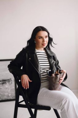 Güzel esmer makyaj sanatçısı beyaz pantolon ve siyah deri ceketli sandalyede oturuyor ve elinde dekoratif kozmetik setiyle bir vazo tutuyor. Güzellik ve moda