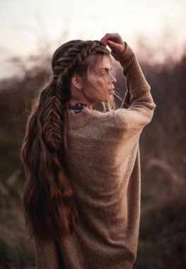 Sonbaharda doğa arka planında şık örgü saç stiliyle poz veren genç ve güzel bir kız. Moda ve güzellik. Güzel sanatlar