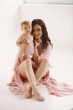 Yeni doğmuş bebeğiyle gülümseyen anne aynı pembe elbiselerle beyaz arka planda stüdyoda poz veriyor. Annenin, 9 aylık bebeğiyle oynadığı pembe gecelikli yakın plan fotoğrafını çek. Metin için alanı kopyala