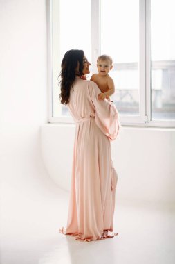 Arka planda, şık pembe elbiseli mutlu bir annenin bebeğini arka planda, stüdyoda tutuşu var. Annelik içeriği, anneler günü konsepti. Arka plan