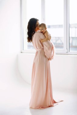 Bebeğini kucağında tutan ve pencerenin yanındaki stüdyoda poz veren güzel bir sabahlık giymiş annenin portresi. Anneler Günü. Mutlu annelik kavramı. Metin için alanı kopyala