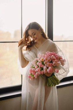Pencere kenarında duran şeffaf beyaz elbiseli ve pembe lale desenli poz veren güzel hamile kadın portresi. Mutlu annelik kavramı. Güzellik ve moda