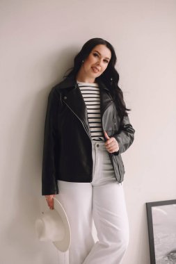 Siyah deri ceketli, beyaz pantolonlu, beyaz şapkalı ve stüdyoda duvarın yanında duran çekici esmer kadın. Güzellik ve moda. Duvar kağıdı ya da baskı dergisi için iyidir.