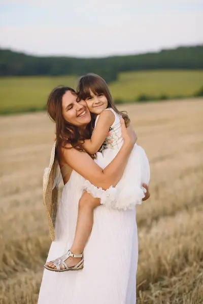Yazlık elbiseli mutlu anne küçük kızını sahada kucaklıyor.