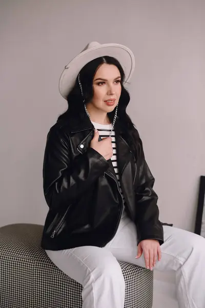 Beyaz şapkalı, siyah deri ceketli ve beyaz pantolonlu güzel esmer kadın stüdyoda oturuyor. Güzellik ve moda. Duvar kağıdı ya da baskı dergisi için iyidir.