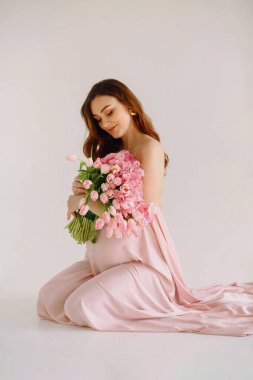 Stüdyoda beyaz arka planda bir buket lale ile pembe kumaşa sarılmış güzel hamile kadın. Hamilelik, annelik, Anneler Günü konsepti. Metin için alanı kopyala
