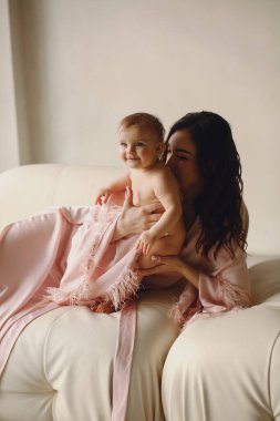 Pembe ipek bornozlu sevimli anne çocuğunun arka tarafını öpüyor. Anne ve bebek. Anne ve bebeğin sevgisi. Mutlu çocukluk kavramı