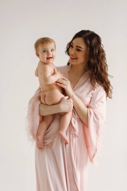 Pembe elbiseli mutlu anne beyaz arka planda poz verirken çıplak bebeğiyle oynuyor. Anne ve kızı. Aile kavramı. Mutlu annelik ve çocukluk kavramı. Anneler Günü