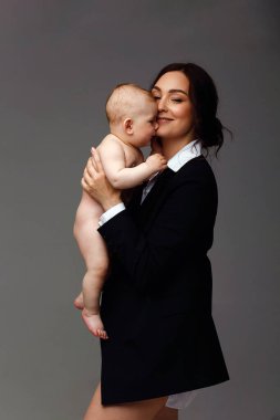 Stüdyo portresi, siyah ceketli ve beyaz gömlekli bir annenin gri arka planda 8 aylık bebeğini büyütmesi. Anne ve bebek. Mutlu annelik kavramı