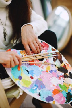 Karmaşık renklerde plastik palet ve boya fırçaları tutan kadın ellerinin yakın çekimi. Sanat stüdyosunda sıradan giyinen kadın sanatçı. Resim atölyesindeki kadın. Yaratıcı etkinlik veya hobi