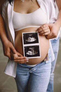 Hamile bir kadın ve partnerinin ultrason fotoğrafını karnına yakın tutarken yakın plan bir fotoğraf. Mutlu ebeveynlik kavramı. Aile sevgisi ve sağlık