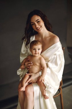 Sandalyeye oturmuş gülümseyen güzel bir kadın. Stüdyoda siyah arka planda poz verirken bebeğini kucağında tutuyor. Mutlu annelik kavramı