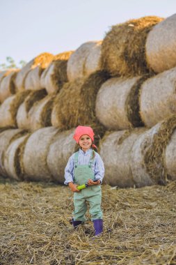 Komik çiftçi kızın dikey çekimi yaz aylarında saman balyalarının arka planında kürekle oynuyor. Kırsalda mutlu bir çocukluk. Mutlu yazlar. Kır hayatı, tarım ve kırsal yaşam tarzı