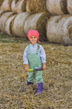 Neşeli çiftçi kız yazın saman balyalarının arka planında kürekle oynuyor. Kırsalda mutlu bir çocukluk. Mutlu yazlar. Kır hayatı, tarım ve kırsal yaşam tarzı