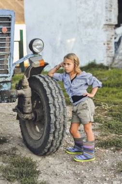 Çiftçi çocuk yazın traktörün yanında duruyor. Kırsalda mutlu bir çocukluk. Mutlu yazlar. Kır hayatı, tarım ve kırsal yaşam tarzı.