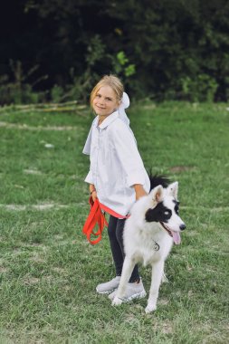 Beyaz tişörtlü, evcil köpeğiyle gezen sarışın bir kızın yaz aylarında doğada dolaştığı dikey bir fotoğraf. Yakutian Laika köpek cinsi. Mutlu çocukluk kavramı