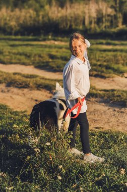 Beyaz tişörtlü gülümseyen küçük bir kızın, yazın köpeğiyle gezerken çekilmiş dikey bir fotoğrafı. Mutlu çocukluk kavramı. Memlekette mutlu yazlar.