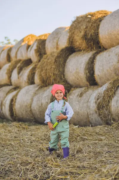 Komik çiftçi kızın dikey çekimi yaz aylarında saman balyalarının arka planında kürekle oynuyor. Kırsalda mutlu bir çocukluk. Uzayı metin için kopyala. Kır hayatı, tarım ve kırsal yaşam tarzı