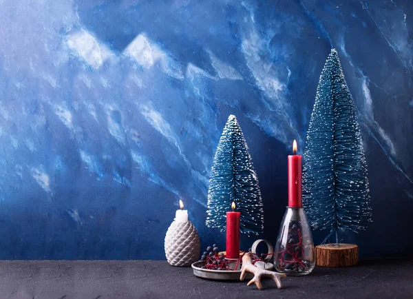 Bright Christmas decorations in red and blue colors. Red burning candles, wild blue berries and blue decorative trees against blue  textured wall. S Still life. Place for text.