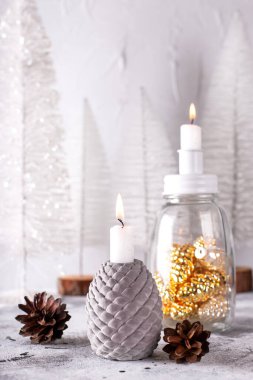 Noel süslemeleri ile kompozisyon. Yanan mumlar, altın dekoratif koni ve doğal kozalaklı cam şamdanlar, gri desenli arka planda beyaz tatil ağaçları. İskandinav minimalist tarzı. Durgun hayat.