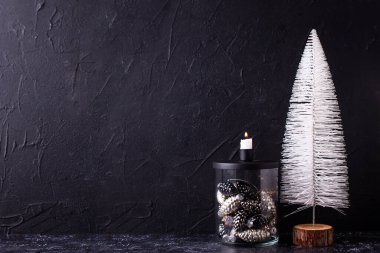   Basit yeni yıl süslemeleri. Şeffaf mum tutacağında dekoratif cam koniler yanan mum ve siyah desenli arka planda beyaz ağaç. İskandinav minimalist tarzı. Durgun hayat. 
