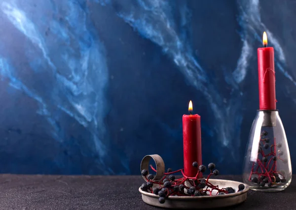 Simple background with burning canles. Candleholders with red burning candles  decorated wild deep blue berries on blue  textured background. Scandinavian minimalistic style. Still life. Place for text.