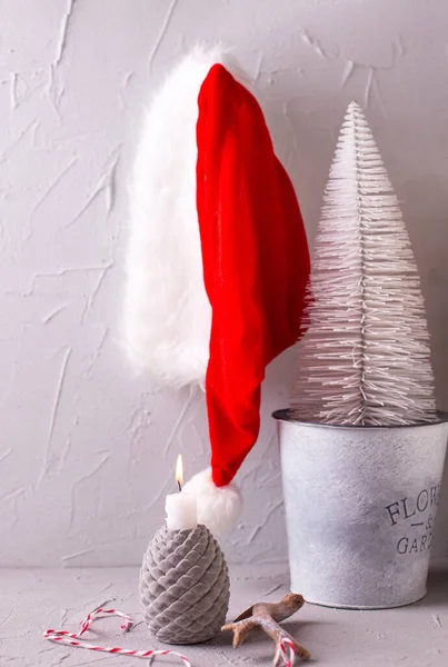 赤で灰色のクリスマスの装飾 白い燃えるキャンドル 白い休日の木 灰色のテクスチャの背景にサンタスの帽子 ノーヴェンミニマリズムスタイル 静物画 — ストック写真