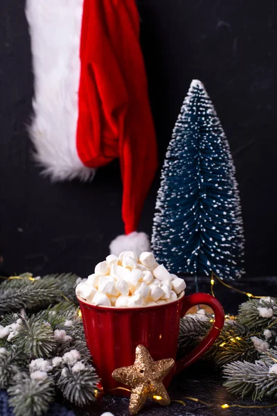 红色的杯子 有热可可和一堆棉花糖 圣诞老人的帽子和蓝色装饰树与黑色纹理背景 圣诞明信片 案文的位置 — 图库照片