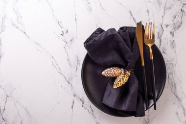 Noel sofrası. Siyah tabak, peçete, altın dekoratif koniler, beyaz mermer arka planda çatal bıçak takımı. Metin için yer.