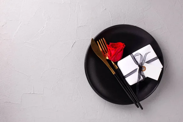 Romantic table setting. Black plate, knife, fork, box with present and empty tag  and red rose flower on textured grey background. Top view. Place fr text. Romantic postcard.